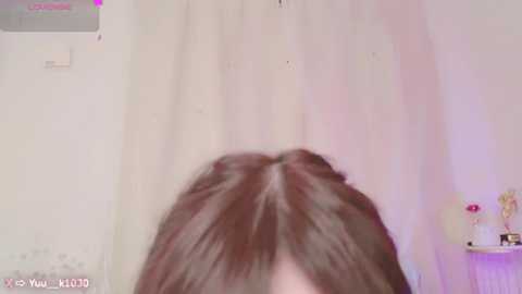 Media: Video of a person with long, straight brown hair, partially visible in a room with a white wall, pink curtains, and a small table with decorative items.