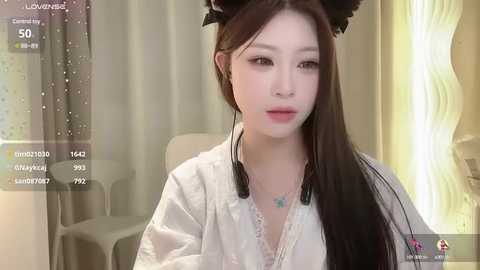 Media: Video of an East Asian woman with long, straight black hair, wearing a white robe, and black cat ear headband, sitting indoors with beige curtains and a lamp.