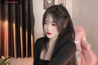 Media: Video of an East Asian woman with fair skin, long black hair, and pointed cat ears, wearing a black top, seated in a pink gaming chair, with a cat figurine in the foreground.