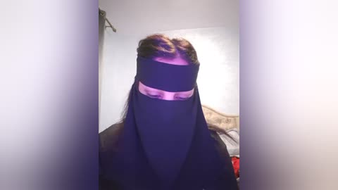 Media: Video of a person wearing a black niqab with a pink bandana covering their hair, standing in a dimly lit room with white walls and a bed in the background.