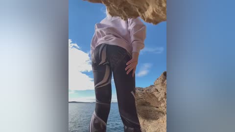 Media: Video of a person in a lavender shirt and black leggings standing on a rocky cliff, facing the ocean under a clear blue sky.