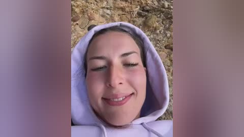 Media: Video of a young woman with light skin and dark hair, smiling, lying on a textured stone ground. She wears a lavender hoodie. The image has a soft, pastel filter.