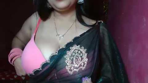 Media: Video of a woman with light skin, wearing a pink bra, black lace shawl, and silver jewelry, leaning against a dark wall.