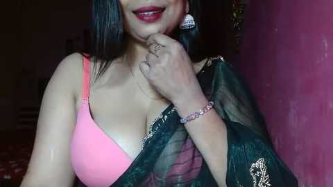 Media: Video of a dark-skinned woman with long black hair, wearing a pink bra and a see-through green sari with gold embroidery. She holds a finger to her lips, smiling. Background features a pink wall and dim lighting.