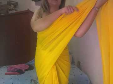 Media: Video of a light-skinned woman with long blonde hair, wearing a yellow sari, standing in a cluttered room with a blue-patterned carpet, wooden dresser, and scattered clothes.