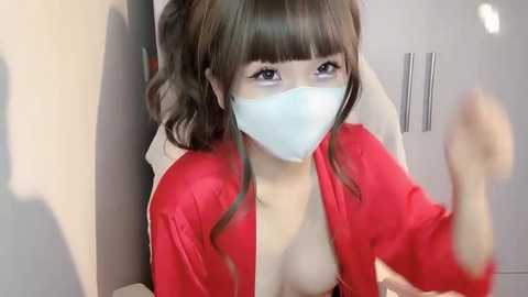 Media: A video of an East Asian woman with long, wavy brown hair and a light complexion, wearing a red satin robe that's open, revealing her bare chest. She's sitting in a white chair with a blue surgical mask covering her mouth and nose.