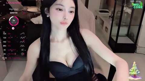 Media: A video of a young Asian woman with long black hair, wearing a black bra, seated in a modern, well-lit room with a glass cabinet and a Christmas tree in the background.