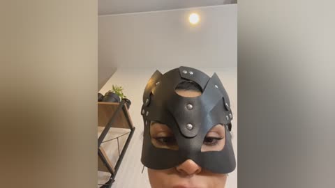 Media: A video of a person wearing a black, geometric leather mask with silver rivets, set against a minimalist, modern interior with a wooden shelf holding succulents and a black geometric plant stand.