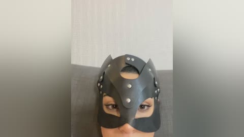 Media: Video of a woman with dark skin wearing a black leather mask with silver studs, covering her eyes and nose, in a neutral-toned room with a textured wall.