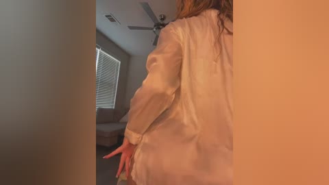 Media: Video of a person with long, wavy brown hair, wearing a sheer, white blouse, standing in a dimly lit room with beige walls and a brown leather couch.