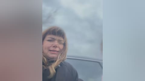 Media: A video captures a woman with long, light brown hair and bangs, smiling slightly, through a car window. The background is blurred, suggesting a cloudy day. She wears a dark jacket.