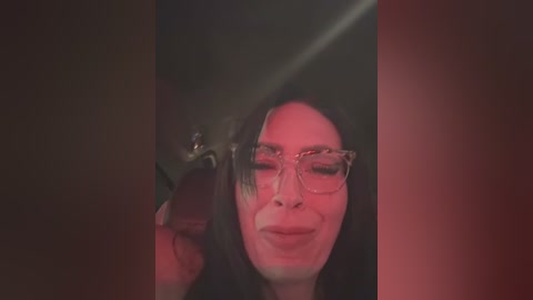 Media: Video of a woman with long dark hair, glasses, and fair skin, smiling in dim light, with red and black color filters overlaying her face.