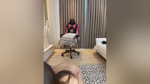 Media: Video of a sparsely furnished room with beige walls, a leopard-print rug, a pink Overwatch gamer chair, and a white bed. A person with a pink bra and pink hair is partially visible, seated on the leopard rug.