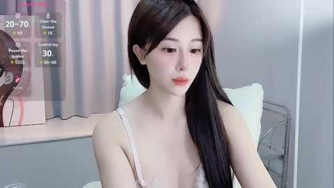 Media: Video of an East Asian woman with long black hair, wearing a white bra, in a hospital room with beige curtains and a bed.