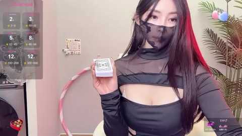 Media: Video of an Asian woman with long black hair, wearing a black mesh top, mask, and red hoop earrings, holding a dollar bill. Background includes a calendar, plant, and wall clock.