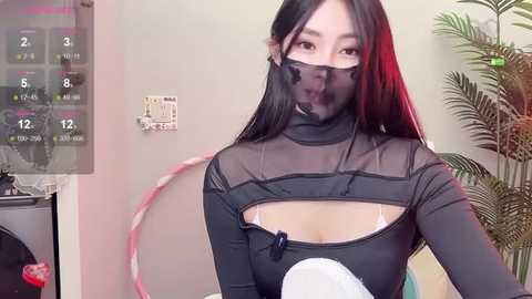 Media: Video of an Asian woman with long black hair, wearing a see-through black bodysuit revealing cleavage, in a room with a calendar, plant, and pink hula hoop.