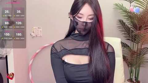 Media: A video of an Asian woman with long black hair wearing a black, see-through, high-necked top and face mask, sitting indoors next to a fake plant.