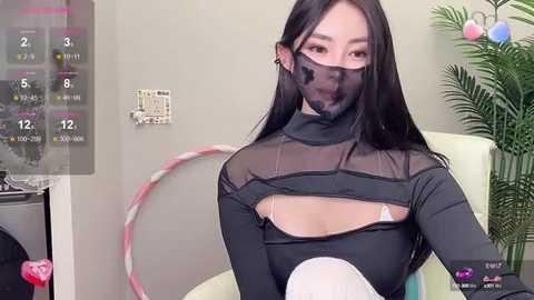 Media: Video of an Asian woman with long black hair, wearing a sheer black mask and a revealing black top, sitting in a modern living room with a hula hoop, plants, and a calendar.