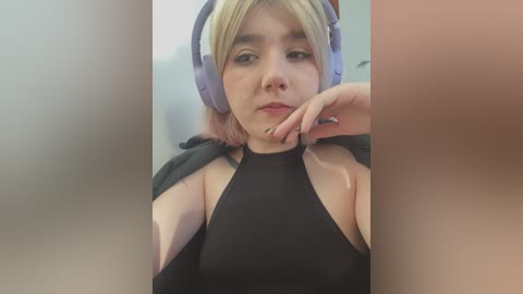 Media: Video of a young woman with pale skin, light blonde hair, and large, over-the-ear headphones, wearing a black halter top. She has a neutral expression, with one hand resting near her mouth.