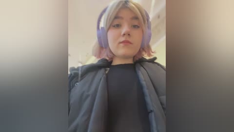 Media: Video of a young person with short, dyed pink hair, wearing purple headphones and a black jacket, blurred background.