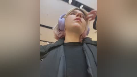 Media: Video of a young woman with light skin and pink hair, wearing purple headphones and a black hoodie, standing in an indoor setting with soft lighting and blurred background.