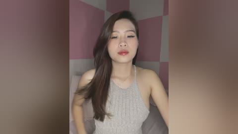 Media: A video of an Asian woman with long, straight, dark brown hair, fair skin, and full lips. She wears a sleeveless, light gray knit dress. The background features pink and white checkered wallpaper.