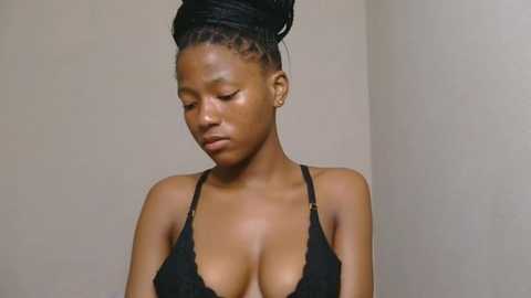 Media: Video of a dark-skinned woman with medium-length braided hair, wearing a black lace bra, standing against a plain beige wall, looking contemplative.