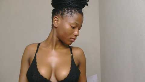 Video of a young, dark-skinned woman with medium-sized breasts, wearing a black lace bra. She has her hair styled in a large, intricately braided bun. The background is plain, off-white walls.