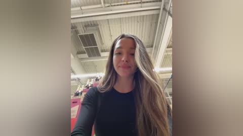 Media: Video of a young woman with long, wavy, light brown hair, wearing a black long-sleeve shirt. She is indoors, in a warehouse with industrial ceiling lights and visible machinery in the background.