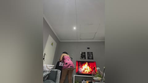 Media: A video shows a woman in a pink dress bending over, facing a lit fireplace in a modern living room with gray walls and a white ceiling.