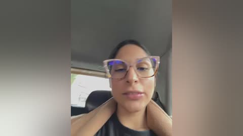 Media: Video of a woman with medium brown skin, wearing large, clear-framed glasses, a black top, and a neutral expression. She is inside a car, with blurred background.