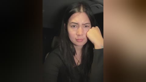 Media: Video of a young woman with long black hair, wearing a black hoodie, resting her head on her hand, appearing sad in a dimly lit car interior.