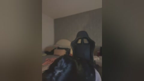 Media: A dimly lit, blurry video of a person wearing a black, possibly leather, bodysuit with a hood, reclining on a bed with disheveled bedding. The room has beige walls and a dark headboard.