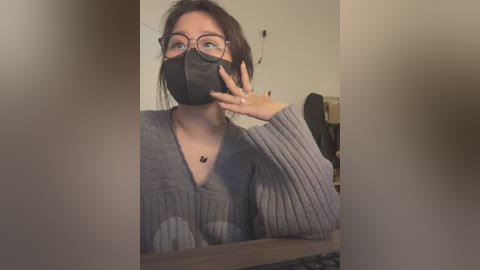 Media: Video of a woman with glasses and a face mask, wearing a gray, ribbed sweater, taking a selfie in a dimly lit room.