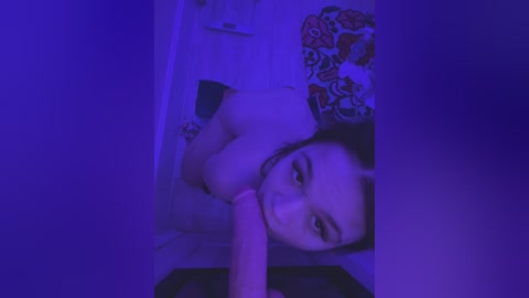 Media: A dimly lit, purple-tinted video of a topless woman with medium-sized breasts, lying on a bed with a floral-patterned sheet, holding a large, erect penis. Her face is partially visible.