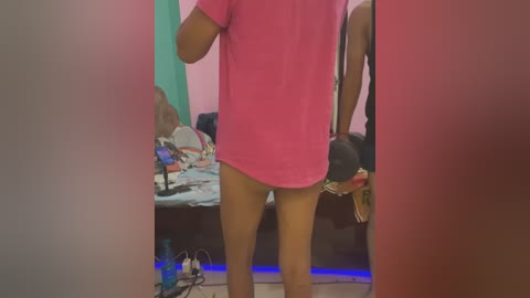 Media: Video of a person wearing a pink t-shirt, standing in a cluttered room with a mirror, green and pink walls, and scattered personal items on the floor.