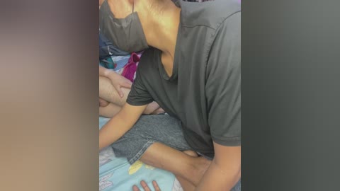 Media: Video of a young, light-skinned male with short hair, wearing a dark grey t-shirt, sitting on a bed with a light blue blanket. His left arm is around a person, partially obscured, with a pink bag in the background.