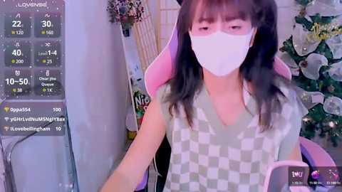 Media: A video of an East Asian woman with long black hair and a white face mask, wearing a checkered shirt, sitting on a pink gaming chair. The background features a Christmas tree and a digital screen displaying streaming stats.