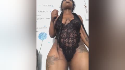 Media: Video of a plus-sized Black woman with dark skin, wearing a black lace bodysuit, standing in front of a white wall adorned with blue floral art and text.