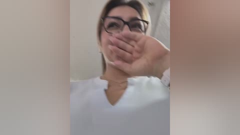 Media: A video captures a young woman with light skin and straight, dark hair, wearing black-rimmed glasses and a white top, covering her mouth with her hand in a bathroom setting.