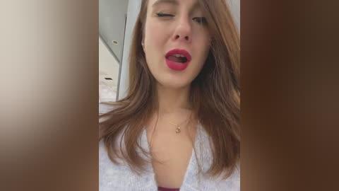 Media: A video of a young woman with fair skin, long brown hair, and red lipstick, wearing a white V-neck sweater, leaning forward, slightly open-mouthed, against a blurred, neutral-toned background.