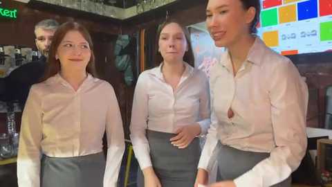 Media: Video of four women in white button-down shirts and dark gray skirts standing in a dimly lit bar, one wearing a small, visible bra.