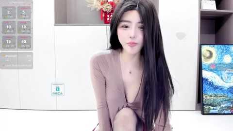 Media: Video of a young East Asian woman with long black hair, wearing a beige V-neck top, seated indoors. Background features a white wall with a temperature gauge, a red flower vase, and a framed painting of The Starry Night by Van Gogh.