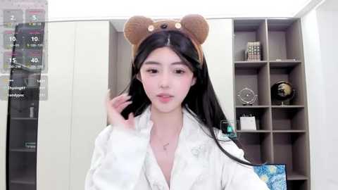 Media: Video of an Asian woman with long black hair wearing a bear-themed headband, white kimono, and makeup, in a modern room with gray shelves.