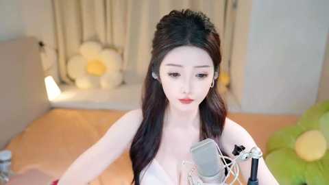 Media: Video of a fair-skinned, long-haired Asian woman with red lipstick, wearing a white dress, singing into a microphone in a bright, cozy room with floral decorations and a green pillow.