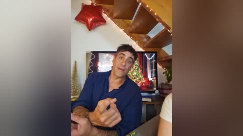 Media: A video captures a man with short dark hair in a blue shirt, gesturing enthusiastically in a cozy living room with a Christmas tree and wooden staircase adorned with fairy lights.