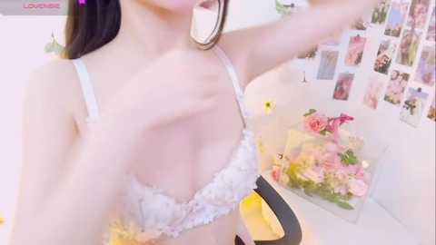 Media: Video of a fair-skinned woman with long dark hair, wearing a lacy white bra, taking a selfie in a brightly lit room with a wall of Polaroid photos and a floral bouquet.