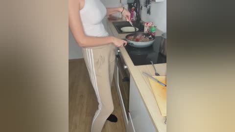 Media: A video of a slim, light-skinned woman in a white tank top and beige Adidas track pants, stirring a pot on a stove in a modern kitchen with wooden floors.