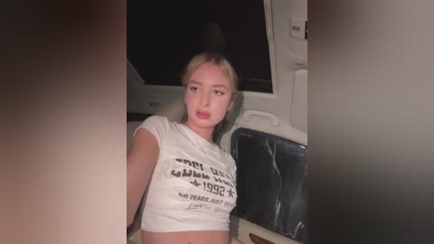 Media: Video of a young woman with fair skin and blonde hair, wearing a white crop top, seated in a car with dim lighting.