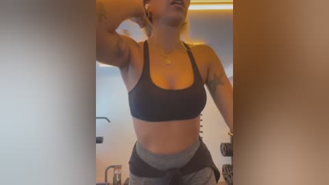 Media: Video of a fit woman with short blonde hair, tattoos, and a black sports bra and grey leggings, posing in a gym.